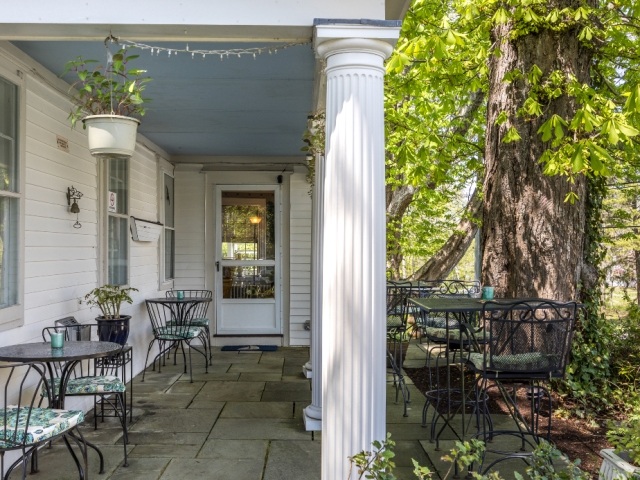 Charming outdoor patio with wrought-iron tables and chairs nestled under a large tree and surrounded by lush greenery at the inn’s entrance.