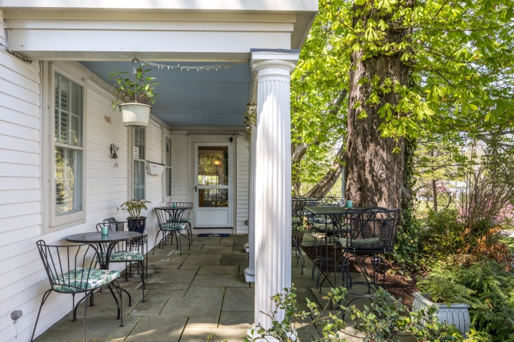 Charming outdoor patio with wrought-iron tables and chairs nestled under a large tree and surrounded by lush greenery at the inn’s entrance.