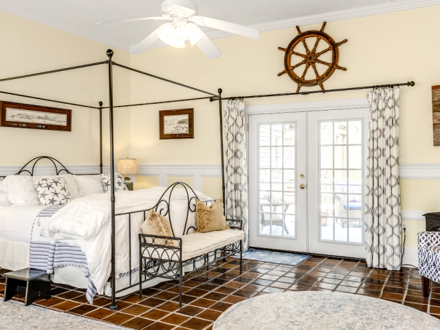 Spacious coastal-themed guest room featuring a four-poster bed, nautical decor, terracotta tile flooring, and French doors leading to a private outdoor area.