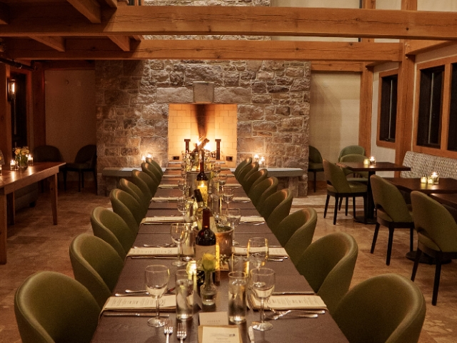 The view of a fireplace over a long table set for dinner under a warmly lit great room with exposed wooden beams