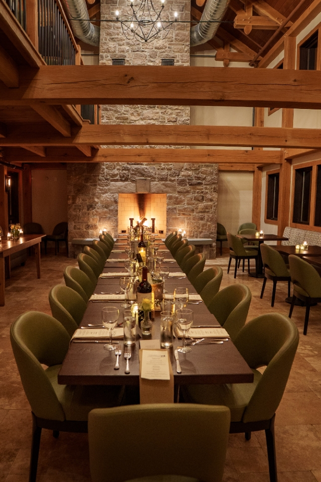 The view of a fireplace over a long table set for dinner under a warmly lit great room with exposed wooden beams