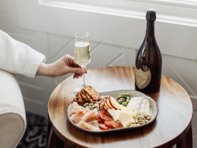 Hand holding a champagne flute beside a Dom Pérignon bottle and a gourmet charcuterie board with meats, cheeses, crackers, and olives.