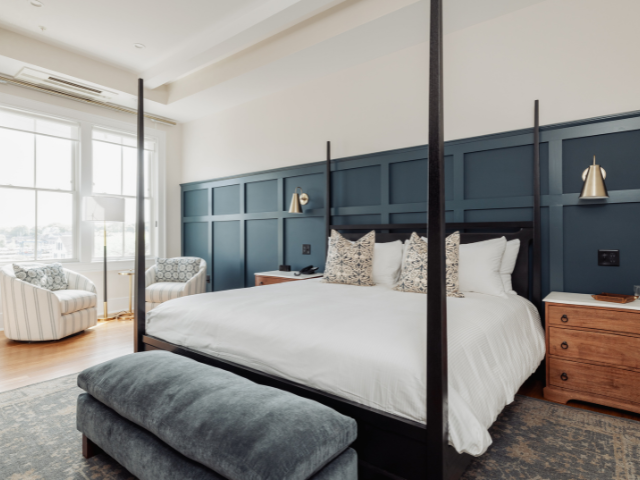 Elegant bedroom featuring a four-poster king bed, blue paneled accent wall, cozy seating, and ample natural light.
