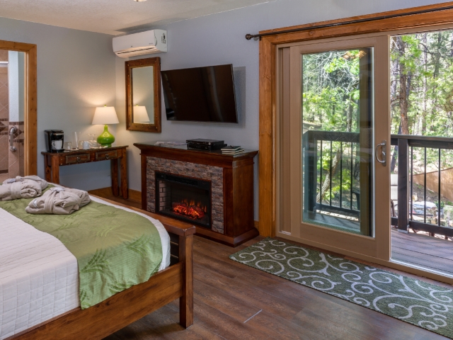 guest room with king bed in front of large tv and fireplace next to a sliding glass door leading out to forest