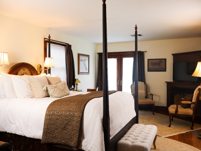 Queen four poster bed with white comforter ad brown blanket, chairs and fireplace