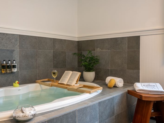 A bathtub with a book, glass of wine on a shelf above the tub, a plant and towels nearby
