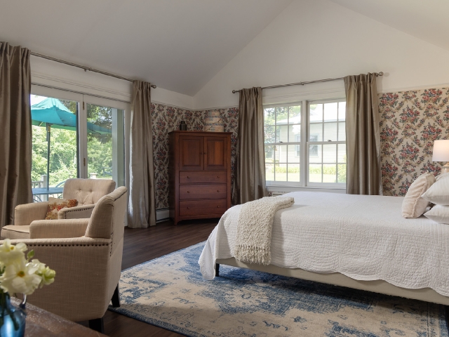 a guest room with a queen bed on a rug and bright windows with the curtains open