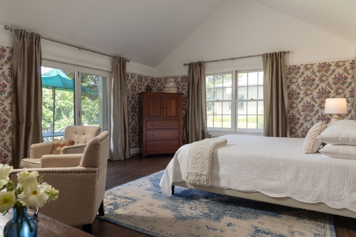 a guest room with a queen bed on a rug and bright windows with the curtains open