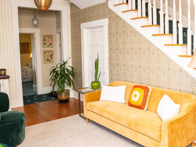 Welcoming foyer with a comfortable sofa, staircase, and greenery, with a doorway leading to a convenient guest refrigerator, iron, and ironing board.