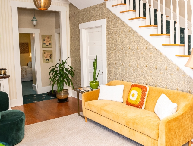 Welcoming foyer with a comfortable sofa, staircase, and greenery, with a doorway leading to a convenient guest refrigerator, iron, and ironing board.