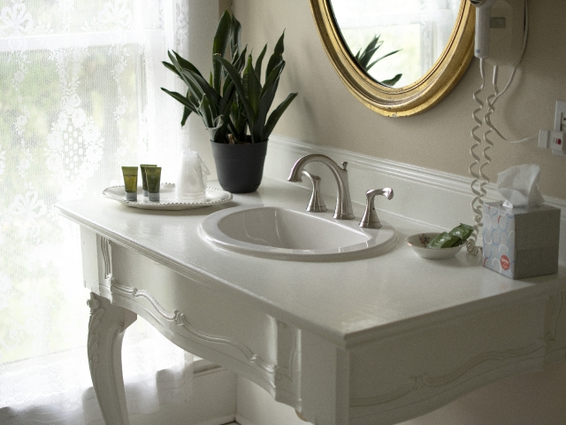 Elegant vanity in the Frederick Suite featuring a bathroom sink set on an antique base, an oval mirror, and a softly lit window framed by lace curtains.