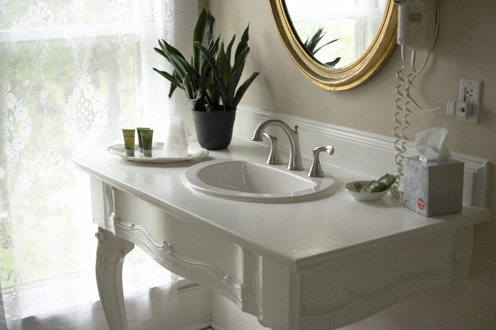 Elegant vanity in the Frederick Suite featuring a bathroom sink set on an antique base, an oval mirror, and a softly lit window framed by lace curtains.