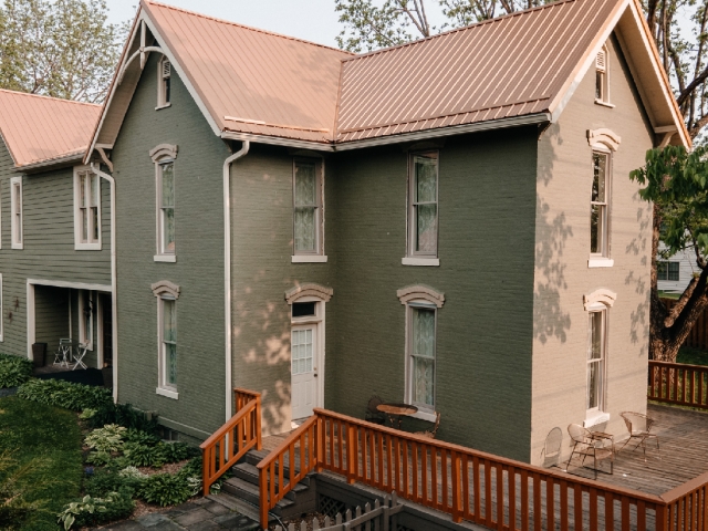 Warm sunlight bathes the front deck of the sage-green Prairie Guest House, highlighting its distinctive copper roof and welcoming country charm.