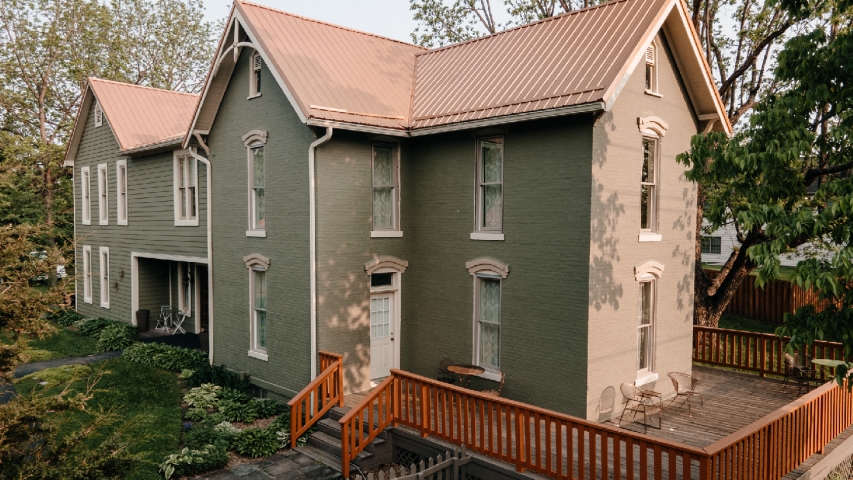 Warm sunlight bathes the front deck of the sage-green Prairie Guest House, highlighting its distinctive copper roof and welcoming country charm.