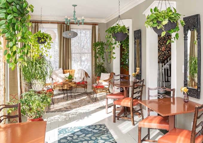 Beautiful, light-filled sunroom with floor-to-ceiling windows on three sides, lush greenery cascading above the tables and chairs, and a peaceful setting where guests gather for breakfast.
