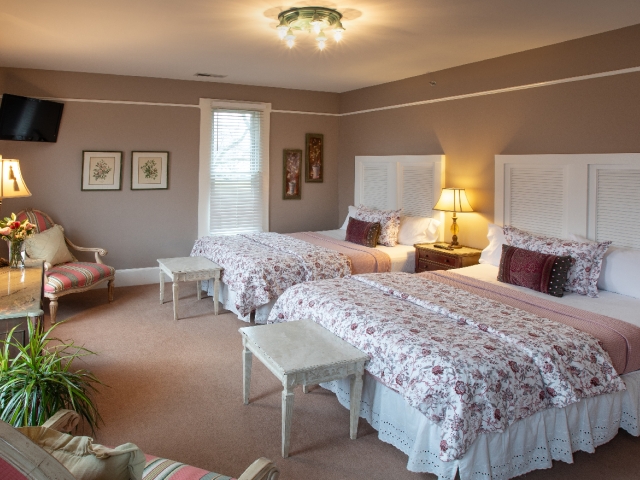 Inviting guest room with two queen beds, cozy wingback chairs, soft natural light, warm bedside lamps, a television, and touches of lush greenery.
