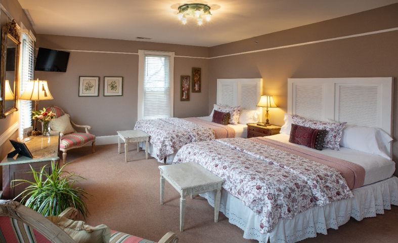 Inviting guest room with two queen beds, cozy wingback chairs, soft natural light, warm bedside lamps, a television, and touches of lush greenery.