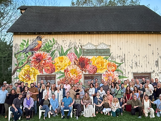 Guests gather for a special birthday celebration and pose for a photo in front of the hand-painted barn mural, surrounded by native trees in our lush green meadow.