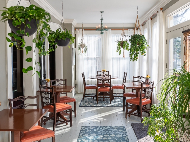 The sunroom glows in the morning light streaming through floor-to-ceiling windows, surrounded by gorgeous greenery and ready to welcome guests for breakfast.
