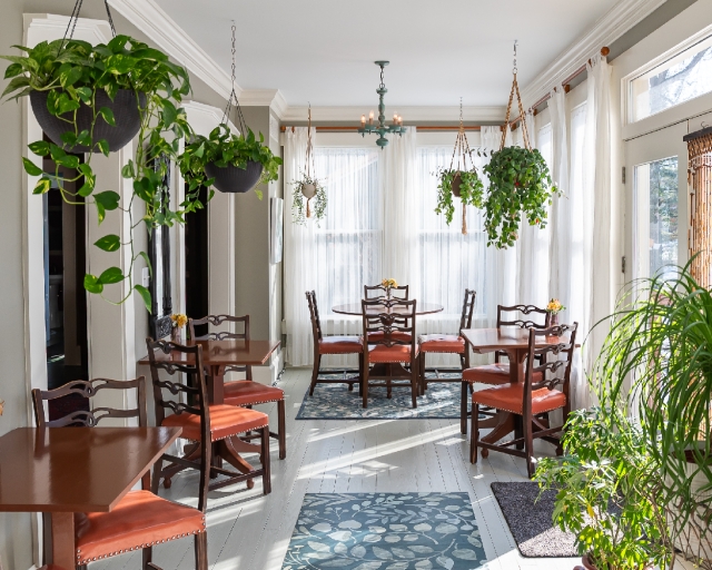 The sunroom glows in the morning light streaming through floor-to-ceiling windows, surrounded by gorgeous greenery and ready to welcome guests for breakfast.
